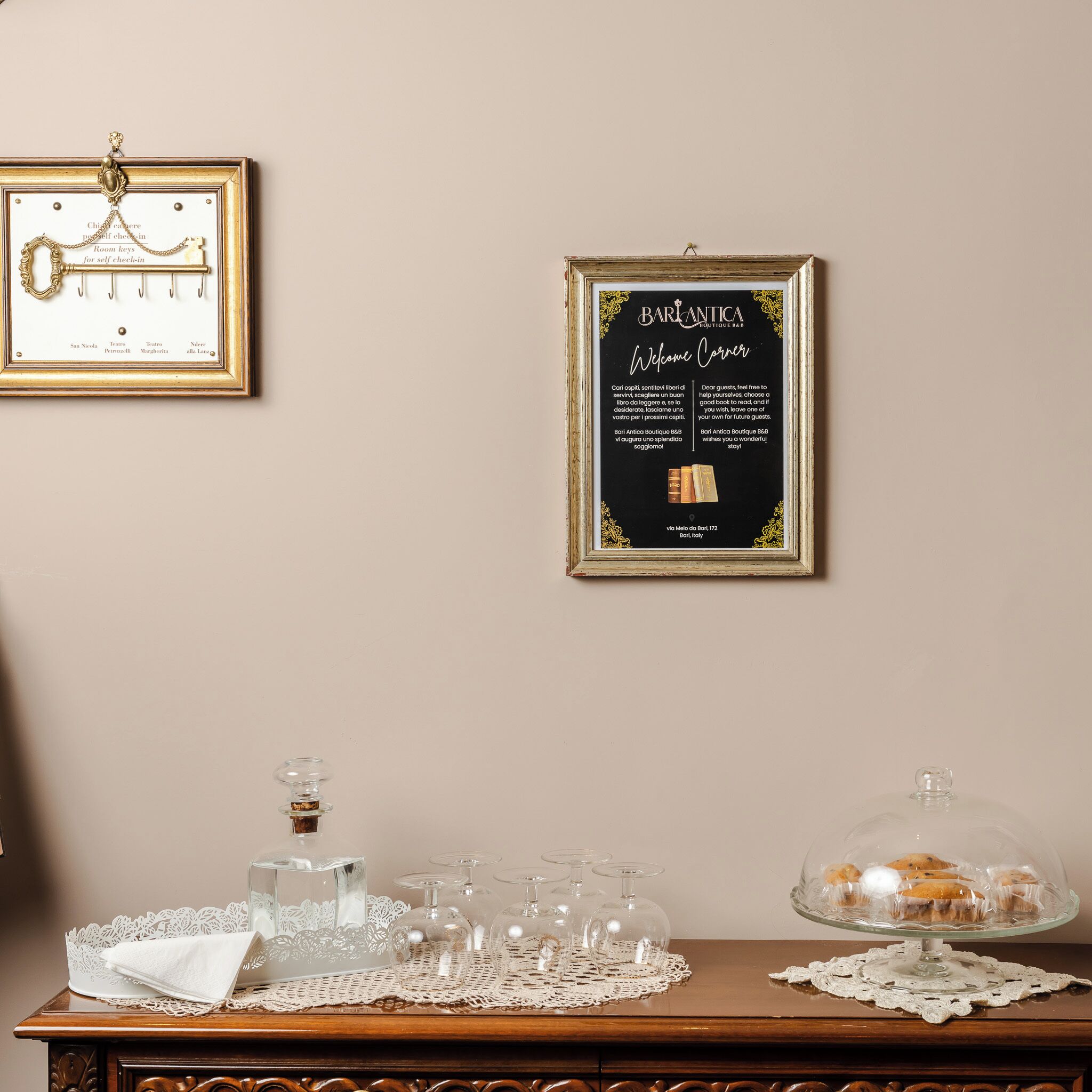 A room with a framed welcome sign, a framed keychain, and a table with a glass cake stand and glasses.