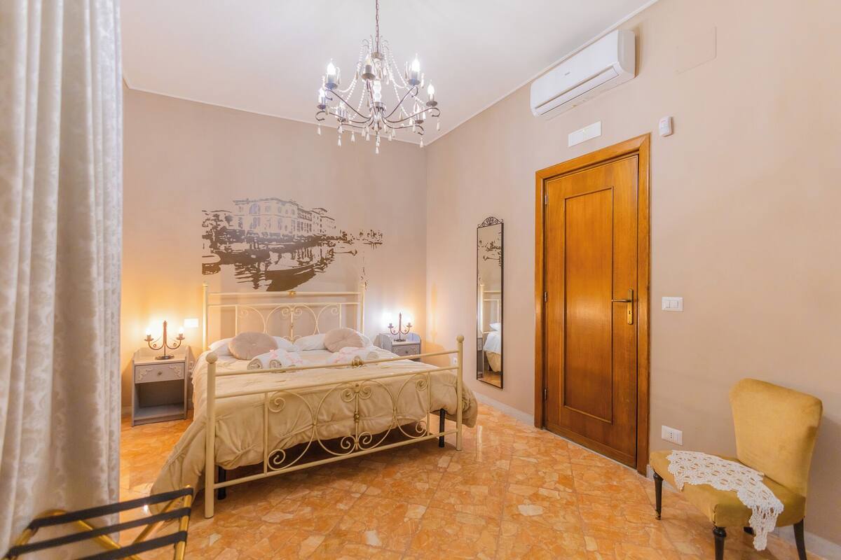 A bedroom with a bed, a chandelier, a wooden door, a chair, and a wall with a cityscape mural.