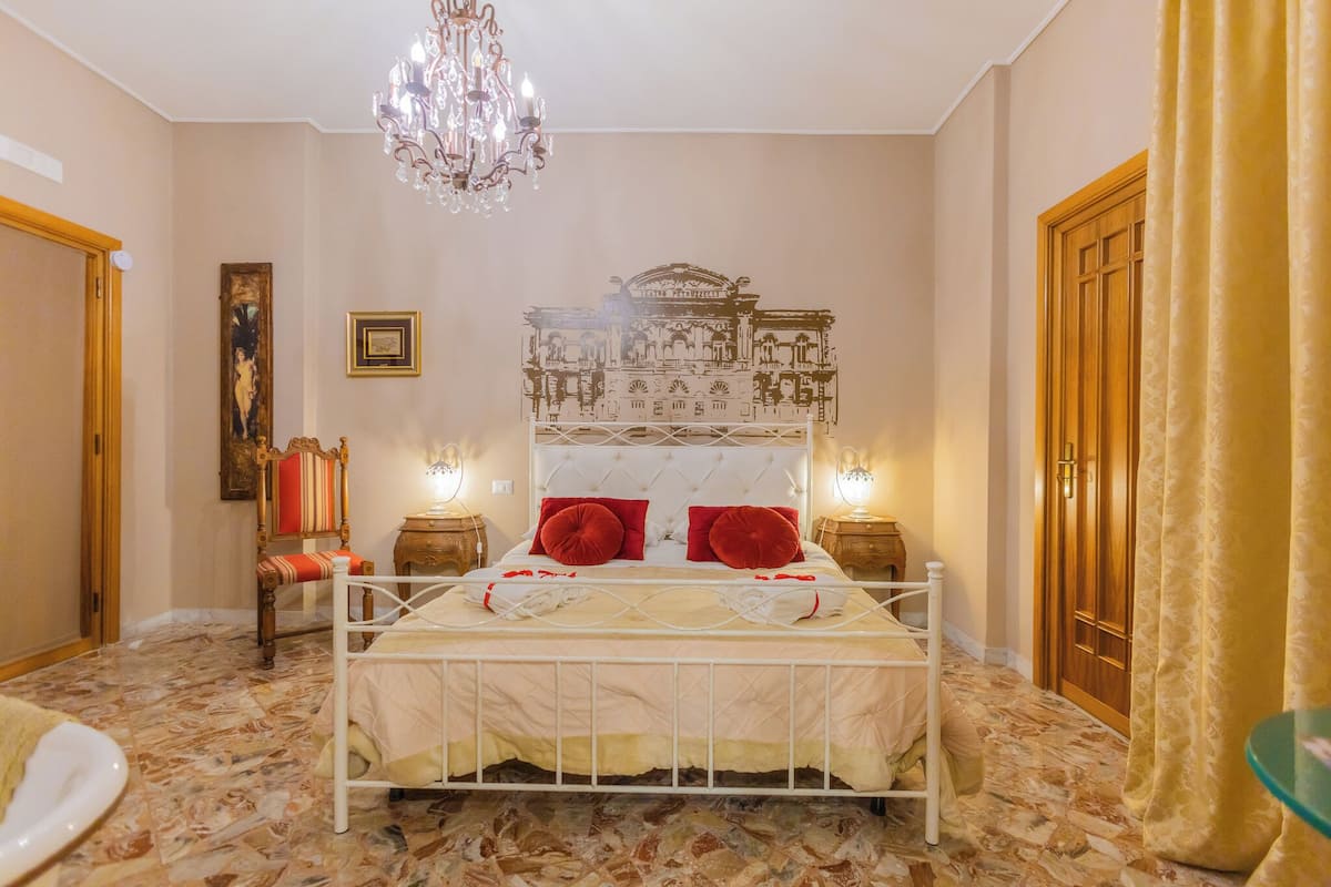 A bedroom with a bed, chandelier, artwork, and a wooden door.