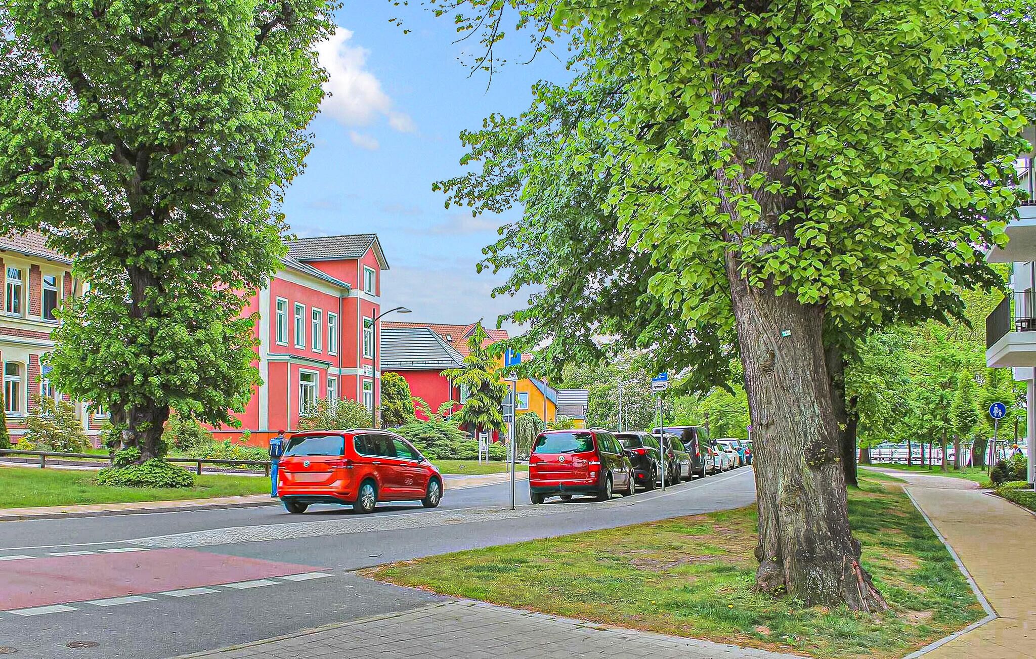 Une voiture rouge roule sur une rue bordée d’arbres, avec des bâtiments en arrière-plan.