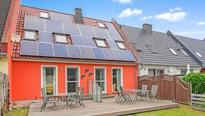 Ein Haus mit roter Fassade, Solaranlage auf dem Dach, einer Holzterrasse mit Gartenmöbeln und einem umzäunten Garten.