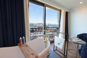 A modern bathroom with a freestanding bathtub, a small table with a towel, and a view of a cityscape through large windows. - Golden Horn Bosphorus Hotel (Istanbul)