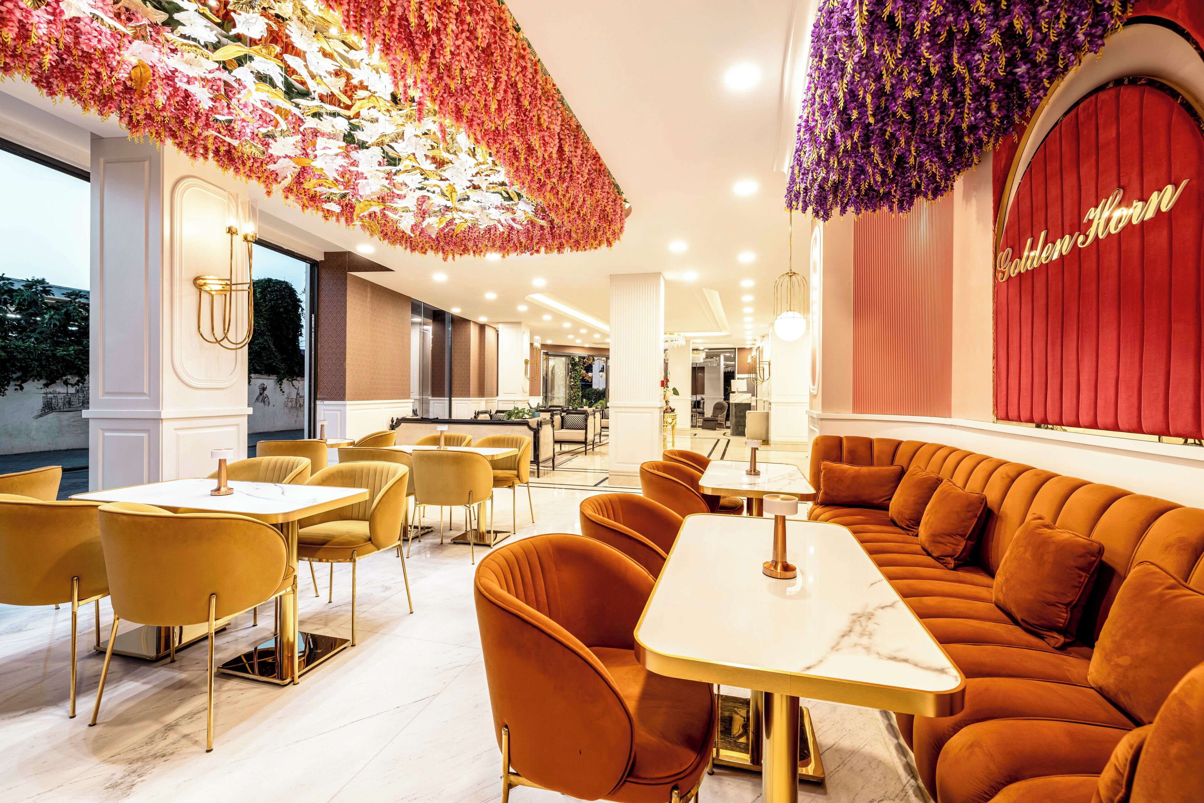 A restaurant with a modern interior, featuring a long orange sofa and yellow chairs, a red curtain with 'Golden Horn' written on it, and a ceiling adorned with hanging floral decorations.
