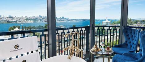 A balcony with a view of a cityscape and the sea, a bathtub, and a blue chair.