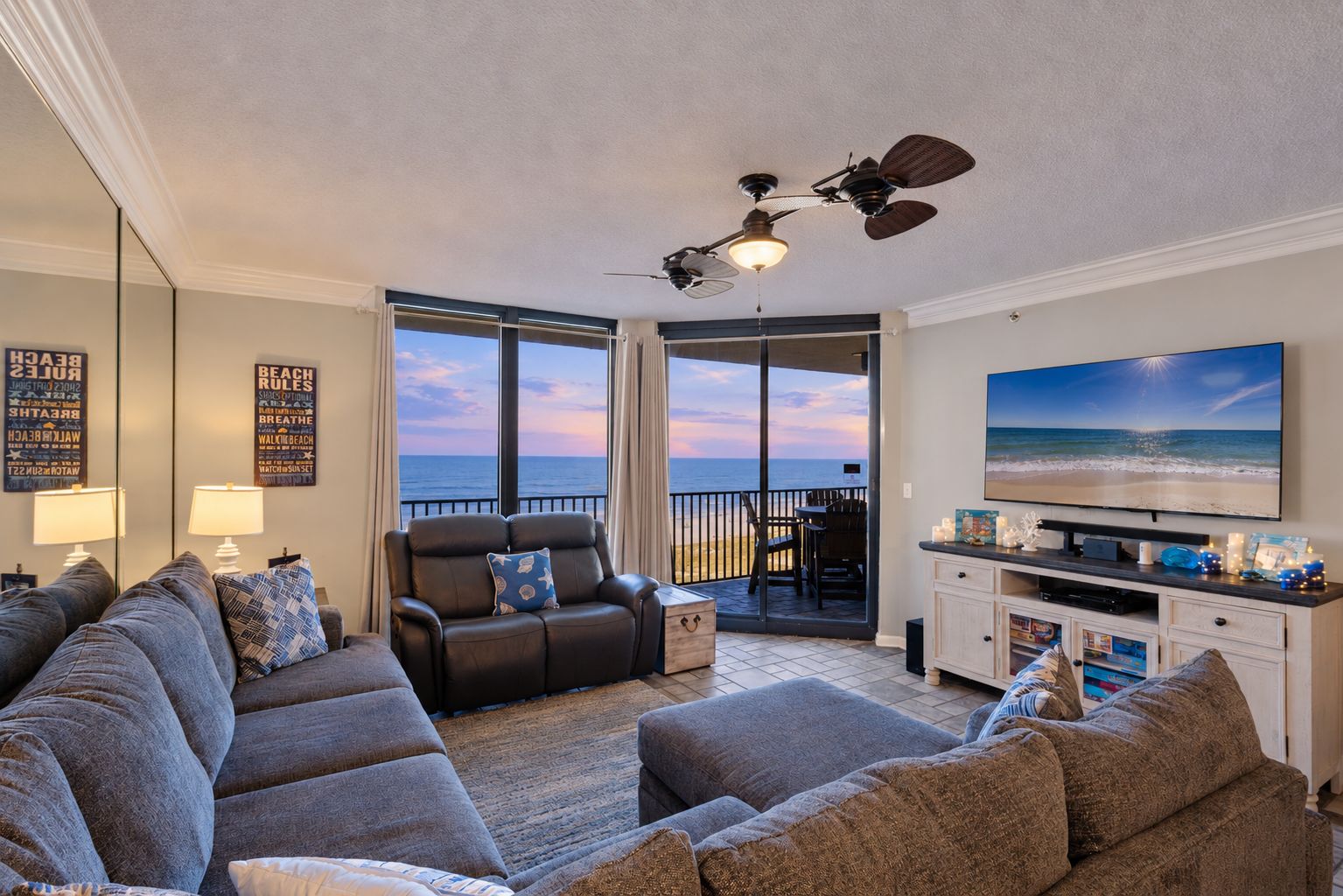 Een woonkamer met een grote televisie, een plafondventilator en uitzicht op het strand.