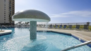 A swimming pool with a water fountain, a poolside lounge area, and a view of a high-rise building in the background. - Phoenix 8 903 | Indoor, Outdoor Pools | Beachfront | Tennis | My Beach Getaways (Orange Beach)
