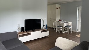 Una sala de estar moderna con un televisor de pantalla plana, un sofá, una mesa de comedor con sillas y una zona de cocina al fondo.