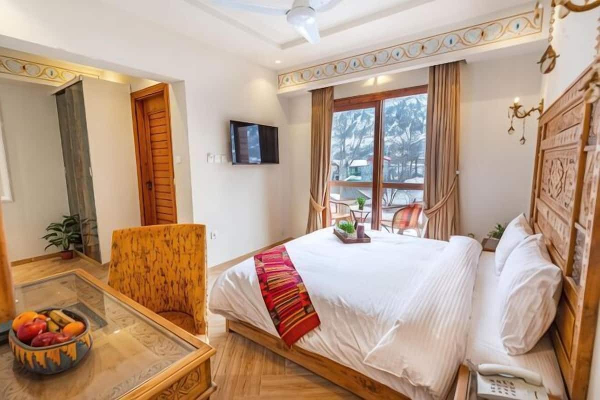 Habitación de hotel con cama, una silla, una mesa con frutero, televisor y ventana con cortinas.