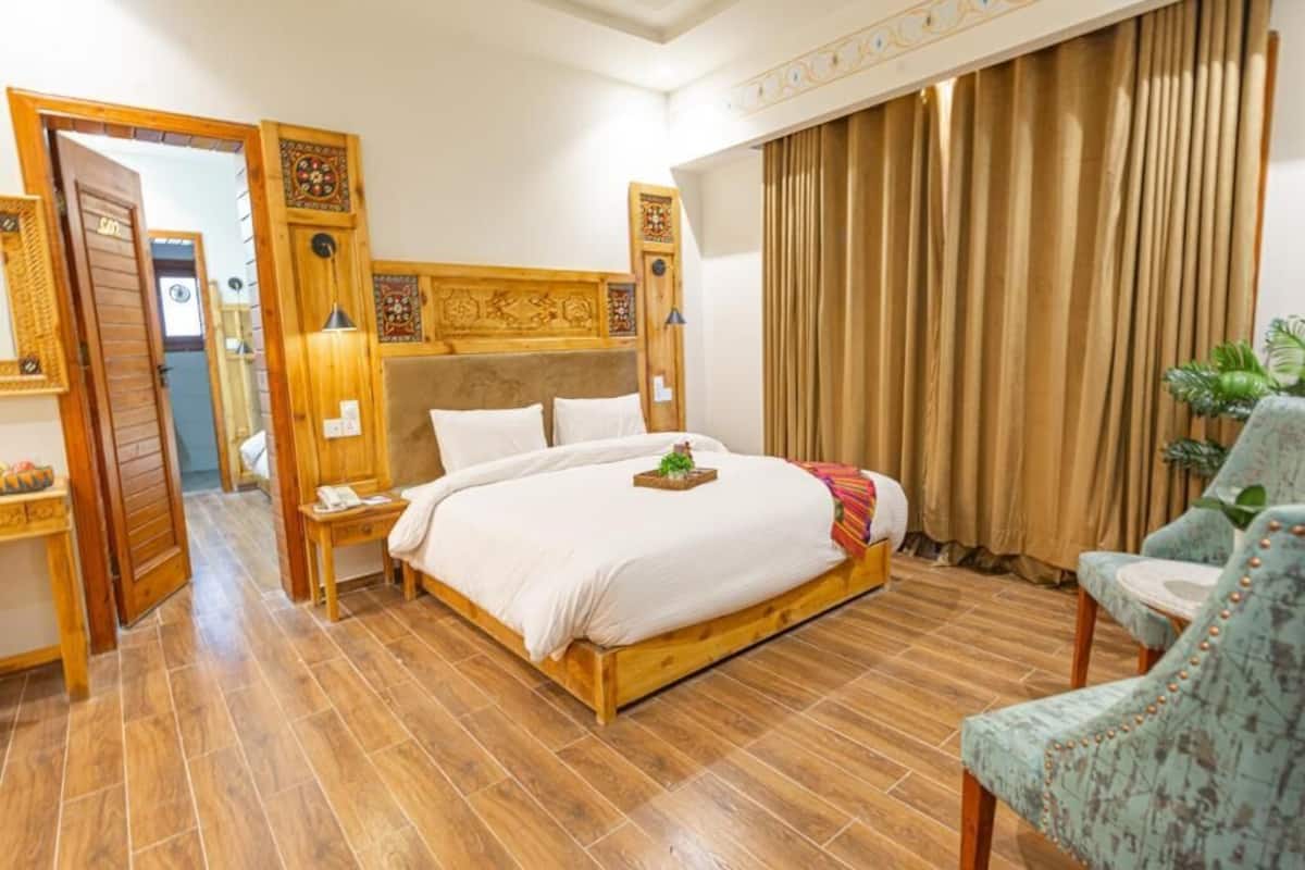 Una habitación de hotel con una cama de madera, una mesita de noche, una silla y un ventanal con cortinas.