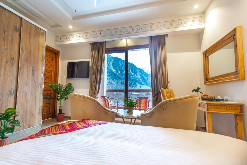 A hotel room with a bed, a desk, two chairs, a TV, a large window with a mountain view, and a wooden wardrobe.