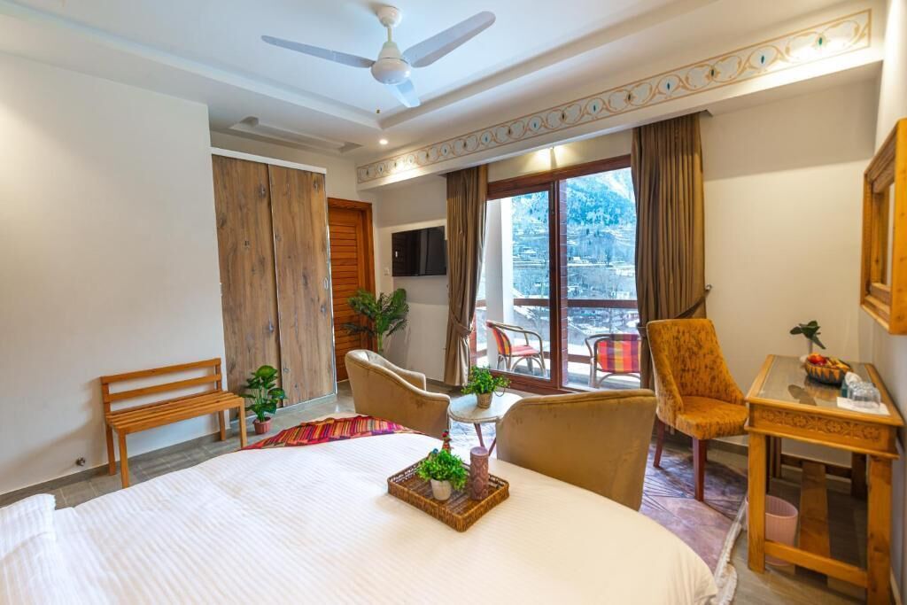 A hotel room with a bed, a ceiling fan, a balcony with chairs, and a view of the mountains.