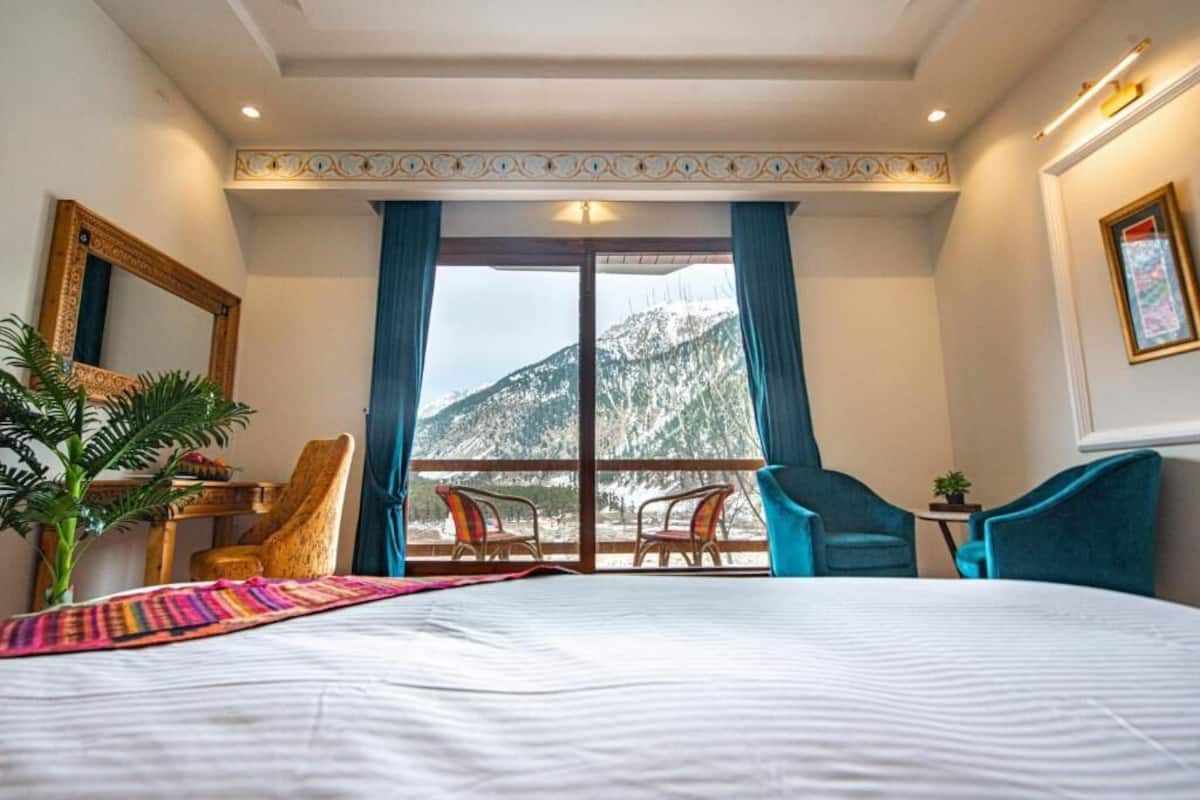 Habitación de hotel con una cama grande, dos sillas, una mesita y vistas a las montañas a través de un ventanal.