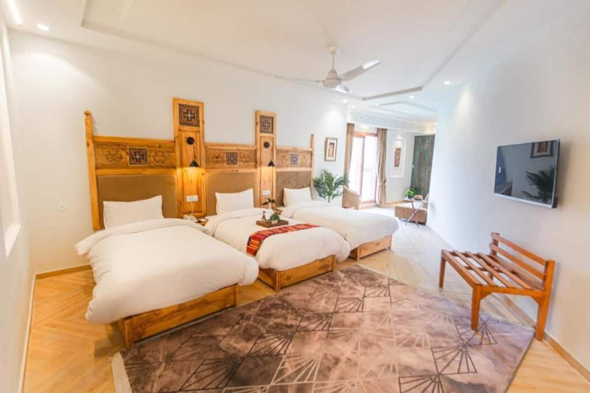 Habitación de hotel con dos camas, cabecera de madera, ventilador de techo, televisor montado en la pared y un banco.