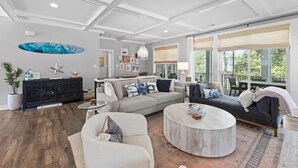 A spacious living room with a large gray sofa, a round coffee table, and a black leather couch. There is a ceiling with recessed lighting and a large window with a view of trees. - 5BR Beauty Near Bethany-Community Pool; Linens (Ocean View)
