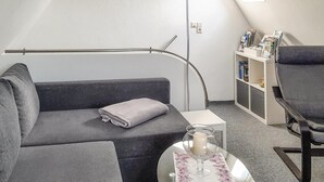 Een moderne kamer met een grijze bank, een klein rond tafeltje met een glazen blad, een kaars en een boekenplank vol tijdschriften.