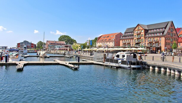 Een jachthaven met aangemeerde boten, een voetpad en gebouwen met winkels.