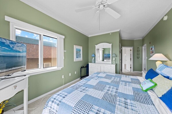A bedroom with a large bed, a ceiling fan, a dresser, a television, and a window with a view of a brick building.