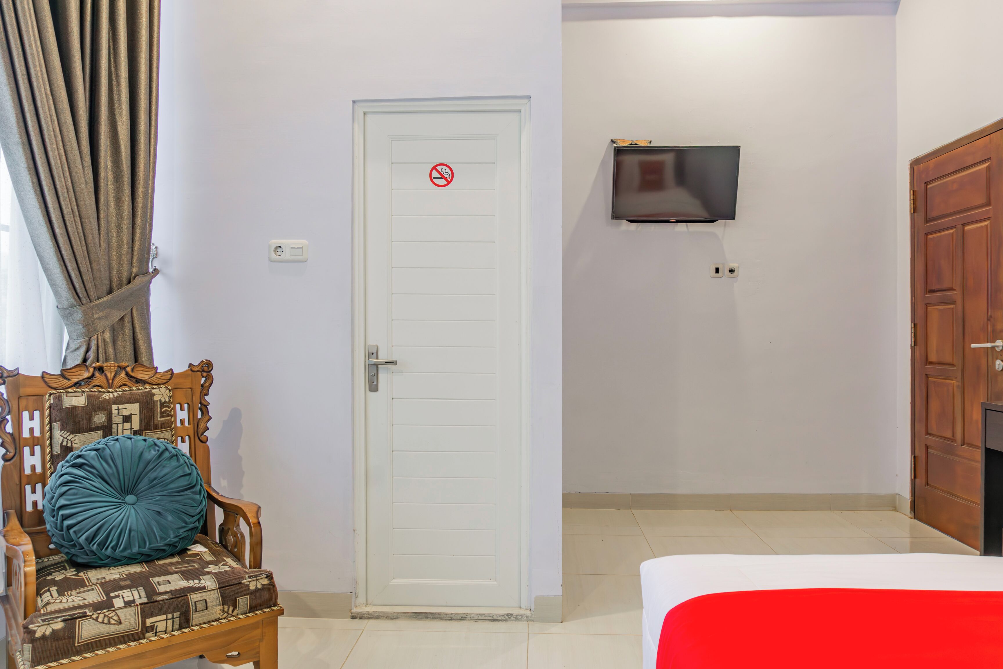 A hotel room with a white door, a television mounted on the wall, a wooden chair with a patterned cushion, and a bed with a red bedspread.