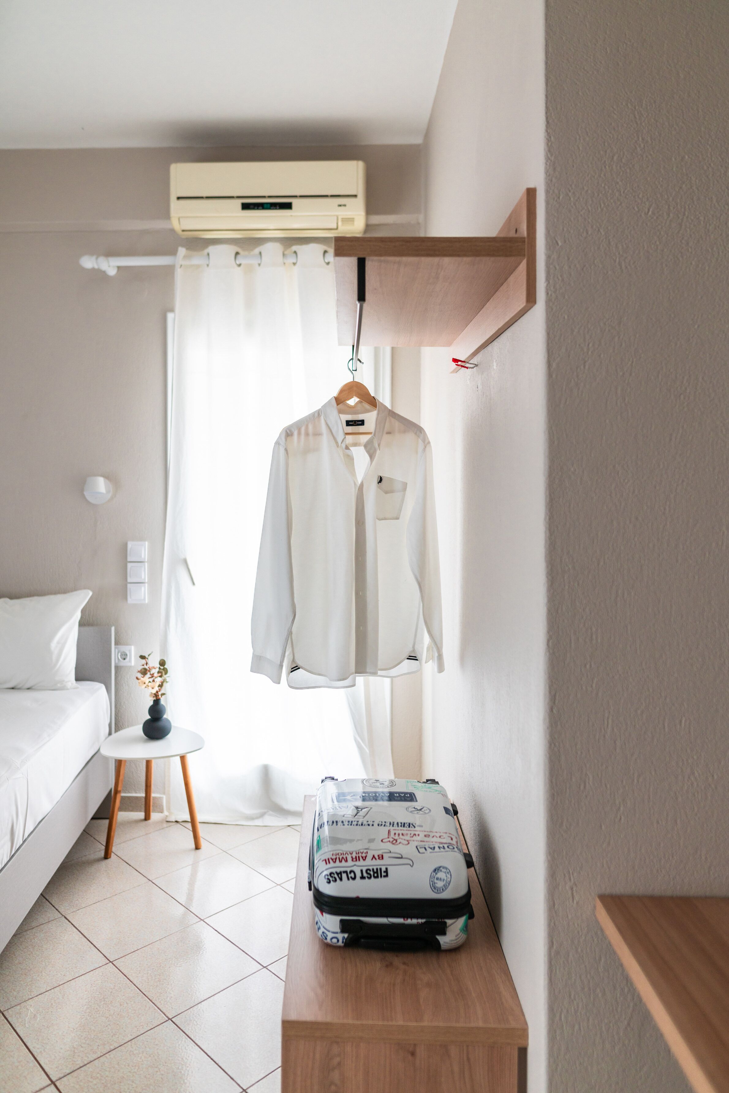Une chambre d’hôtel avec un lit, une petite table sur laquelle se trouve une valise, une chemise suspendue et un appareil de climatisation.
