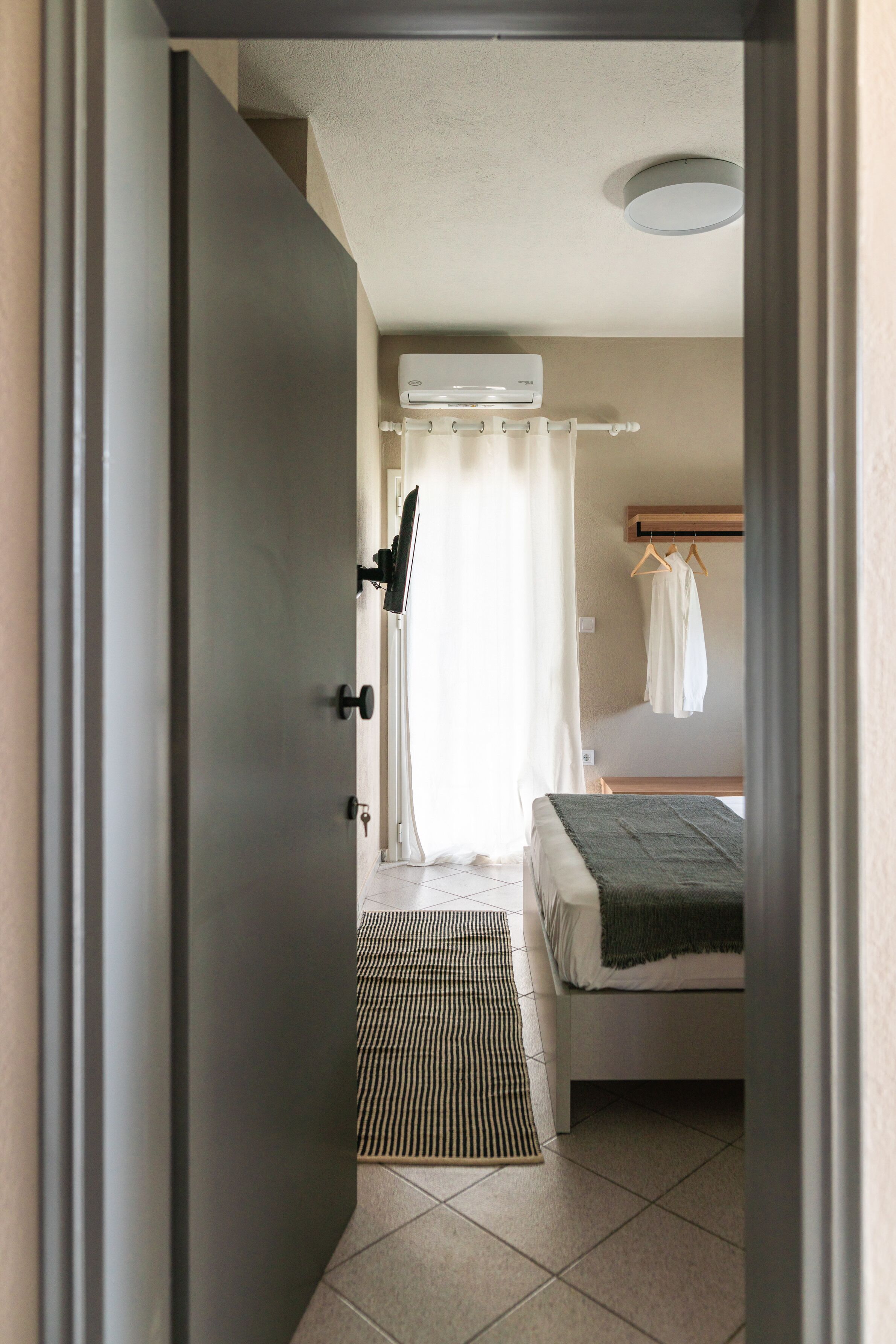 A room with a bed, a bedside table, a hanging light, a window with curtains, and a wall-mounted air conditioner.