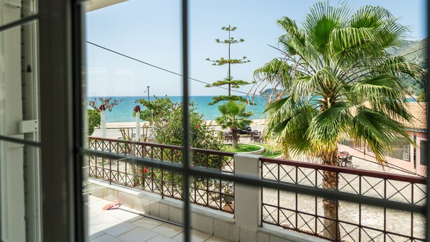 A balcony with a view of the sea, palm trees, and a building with a red roof. - Blue Hug Apartments  (Igoumenitsa)