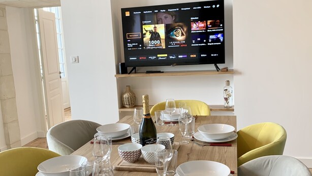 A dining table set with plates, glasses, and cutlery, a bottle of champagne, and a TV mounted on the wall displaying a streaming service interface. - Entre Loire et Château (Amboise)