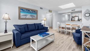 A modern living room with a blue sofa, a white coffee table, and a painting of a beach scene. - Pelicans Landing Unit 409 - Ocean view (Myrtle Beach)