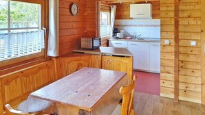 En inredning med träpanel, ett matbord, stolar, ett köksområde med vita skåp och ett fönster med persienner.