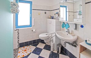 Een badkamer met toilet, wastafel en spiegel. Er is een handdoekhouder met handdoeken, een klein schapje met toiletartikelen en een vloermat met dessin.
