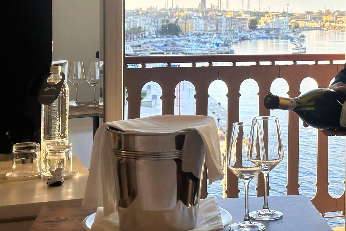 A table with a bottle of water, glasses, and a metal container, overlooking a marina with boats and buildings.