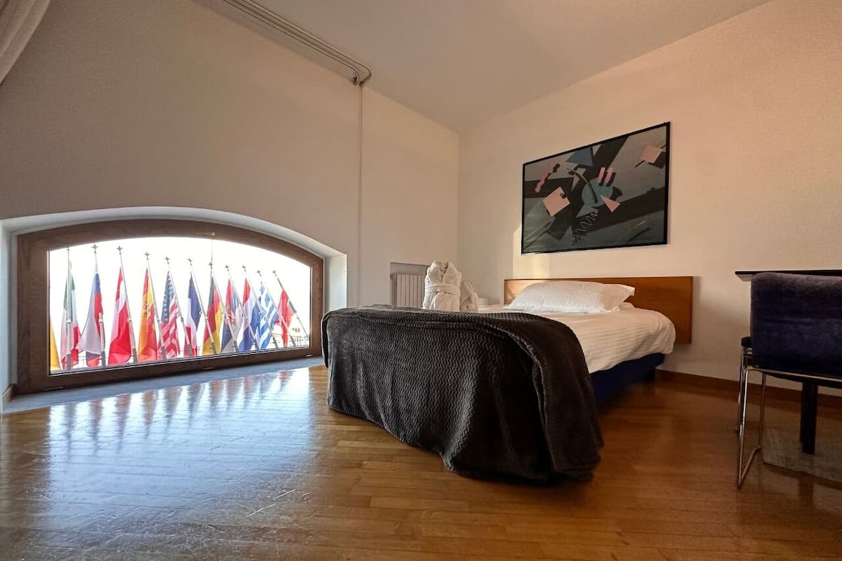 A hotel room with a large window overlooking a row of international flags, a bed with a white comforter, a dark blanket, and a painting on the wall.
