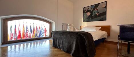 A hotel room with a large window overlooking a row of international flags, a bed with a white comforter, a dark blanket, and a painting on the wall.