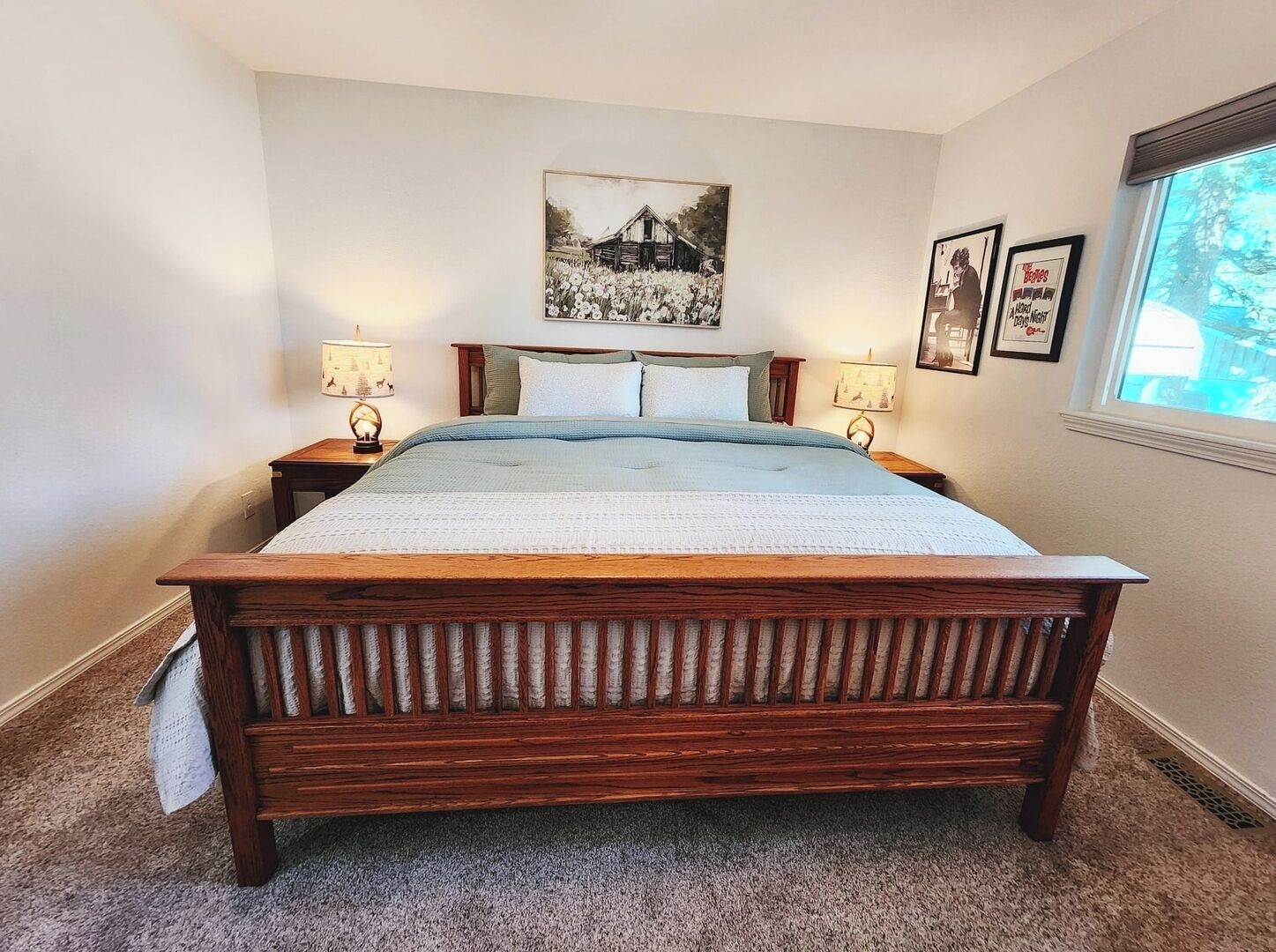Een slaapkamer met een houten bed, twee nachtkastjes met lampen, een groot schilderij in een lijst aan de muur en een raam met uitzicht op bomen.
