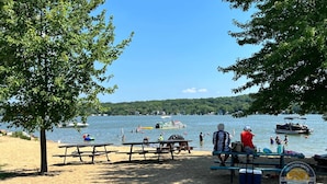 A lakeside picnic area with benches and tables, boats on the water, and a forested hill in the background. - Exclusive Lake Como Escape with Bar and Pool Table (Lake Geneva)