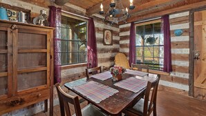 Interior rústico de una cabaña de madera con una mesa de comedor, sillas, una araña y una ventana con cortinas.