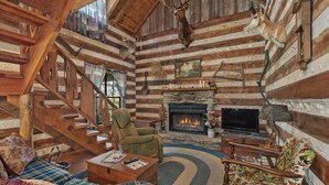 Interior de una cabaña de troncos con chimenea, televisor, sofá, sillón y escalera.