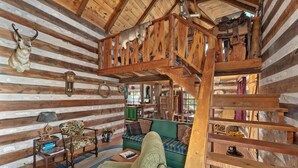 Interior rústico de una cabaña de troncos con un altillo, paredes de madera, un sofá verde, un sillón estampado y una escalera de madera.