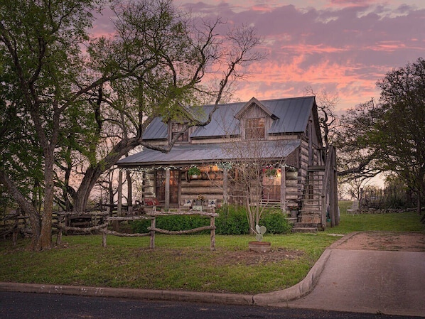 A rustic log cabin with a porch, surrounded by trees and a well-maintained lawn.
