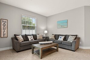 A living room with two grey sofas, a wooden coffee table, a window with blinds, a lamp, and a framed picture on the wall. - 5BR family townhome near the beach w/ pool (Indian Harbour Beach)