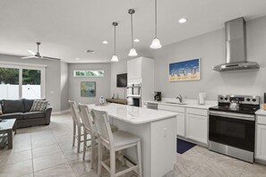 Una cocina moderna con armarios blancos, una isla central con taburetes de bar, electrodomésticos de acero inoxidable e iluminación colgante.