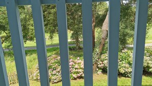 Une vue à travers une clôture blanche, donnant sur un jardin avec de l’herbe, des arbres et des fleurs roses.