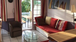 Un salon avec une table basse en verre, un canapé avec des coussins à rayures, un fauteuil et une table à manger avec des chaises. Un tableau représentant des planches de surf est accroché au mur, et il y a une fenêtre avec des rideaux.