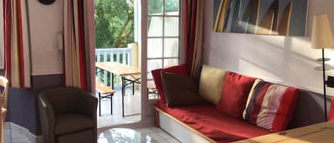 A living room with a glass coffee table, a sofa with striped cushions, a chair, and a dining table with chairs. There is a painting of surfboards on the wall, and a window with curtains.