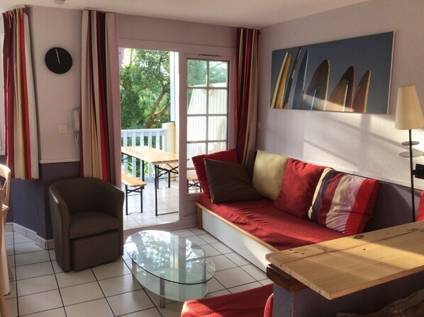 Un salón con una mesa de centro de vidrio, un sofá con cojines a rayas, una silla y una mesa de comedor con sillas. Hay un cuadro de tablas de surf en la pared, y una ventana con cortinas.