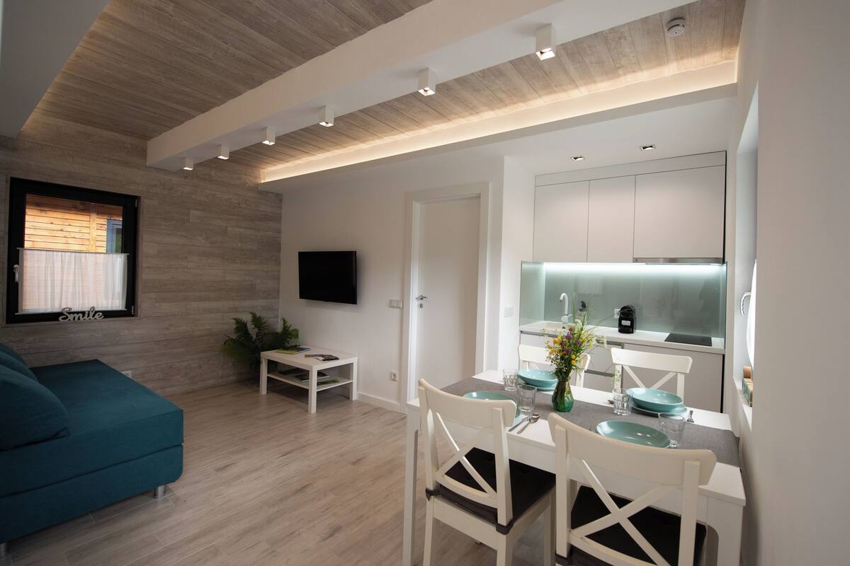A modern living room with a teal sofa, a dining area with white chairs, a kitchen with a glass backsplash, and a flat-screen TV mounted on the wall.