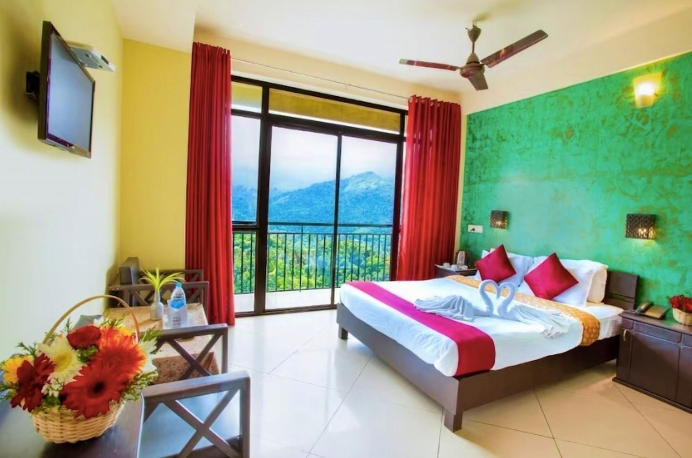 Habitación de hotel con cama, escritorio, silla, televisión, balcón y ventilador de techo, con vistas a las montañas.