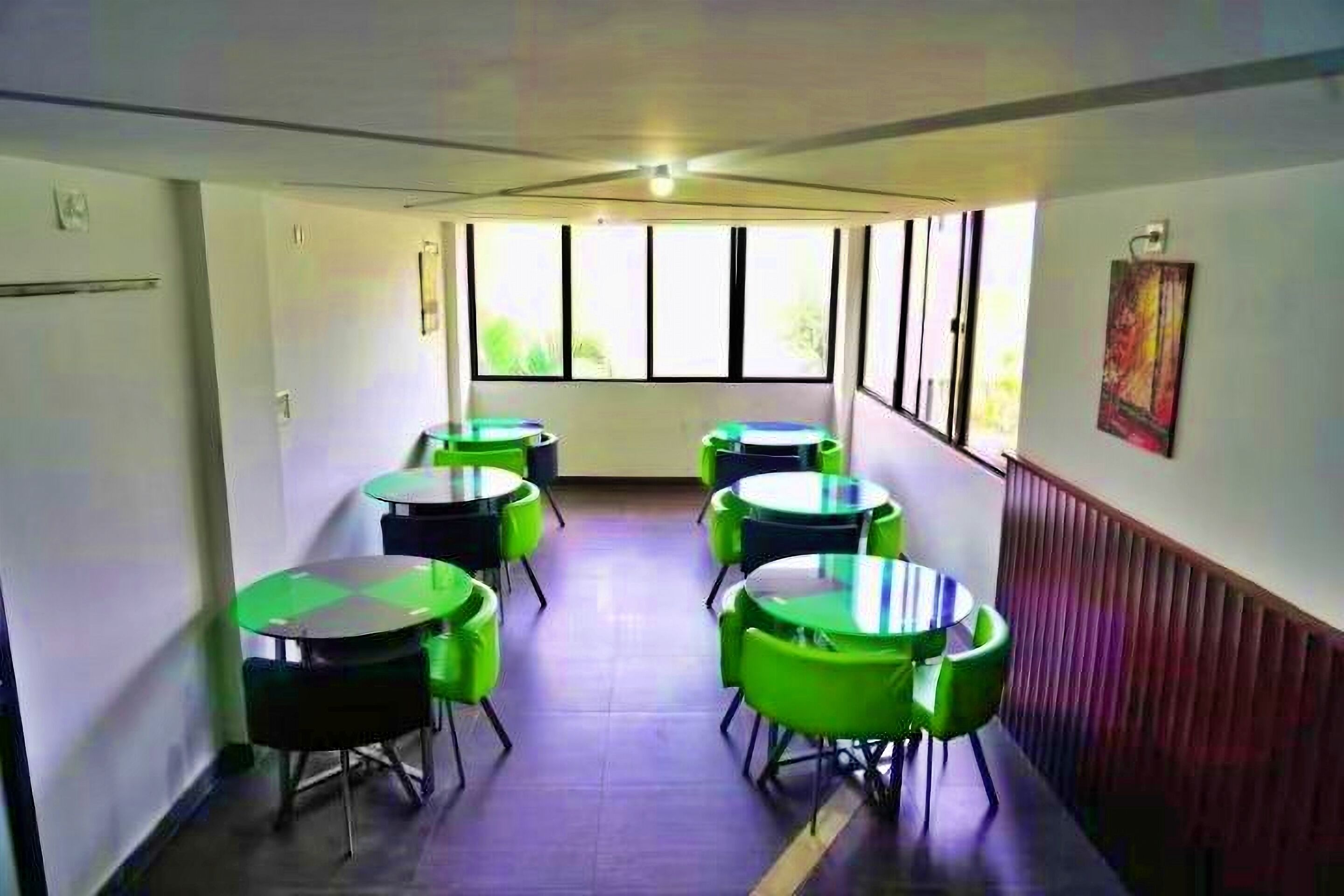 A room with multiple round tables and chairs, large windows with blinds, and a radiator on the wall.