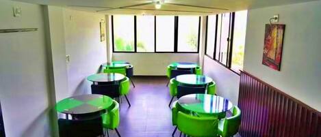 A room with multiple round tables and chairs, large windows with blinds, and a radiator on the wall.