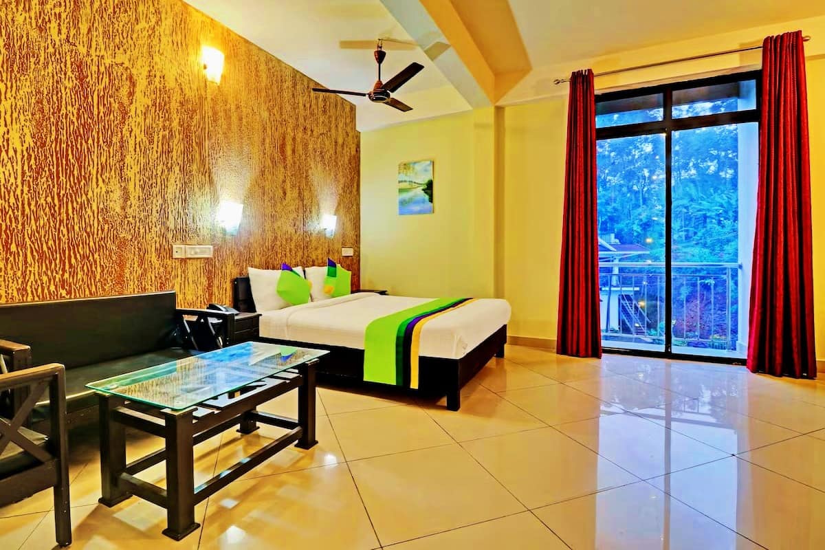 A hotel room with a bed, a sofa, a glass-top table, a ceiling fan, and a large window with curtains.