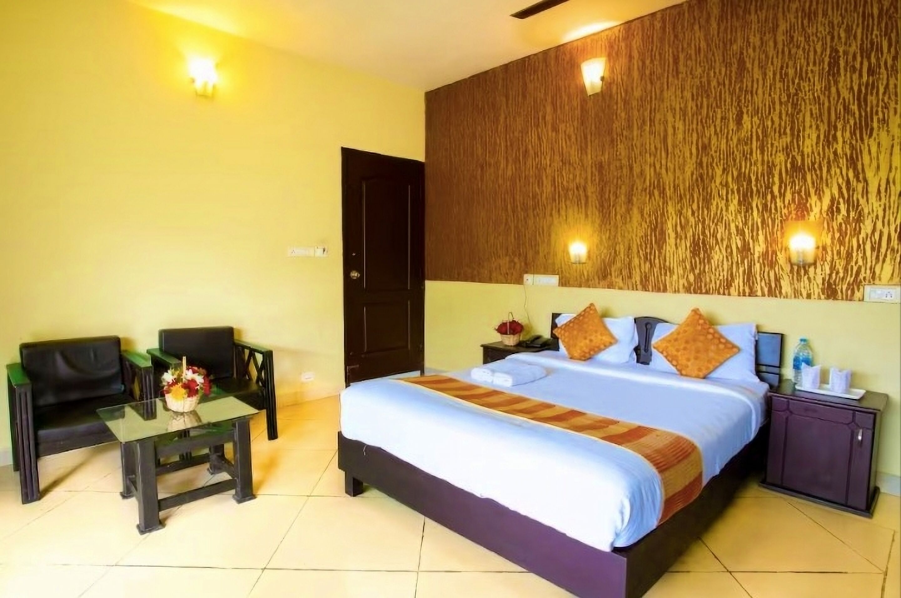 A hotel room with a bed, a small table with a vase of flowers, a sofa, a nightstand, and a wall-mounted light fixture.